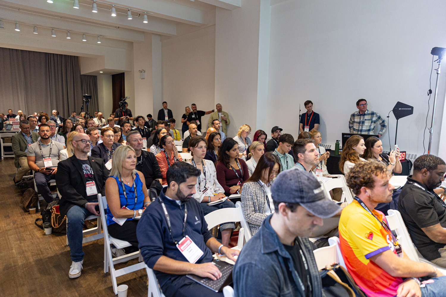 A room of seated attendees with lanyards, some with laptops, facing the front of the room.