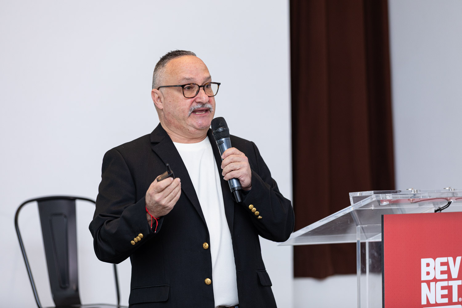 A man in glasses, a black blazer and a white t-shirt speaks into a microphone next to a BevNET Live podium.