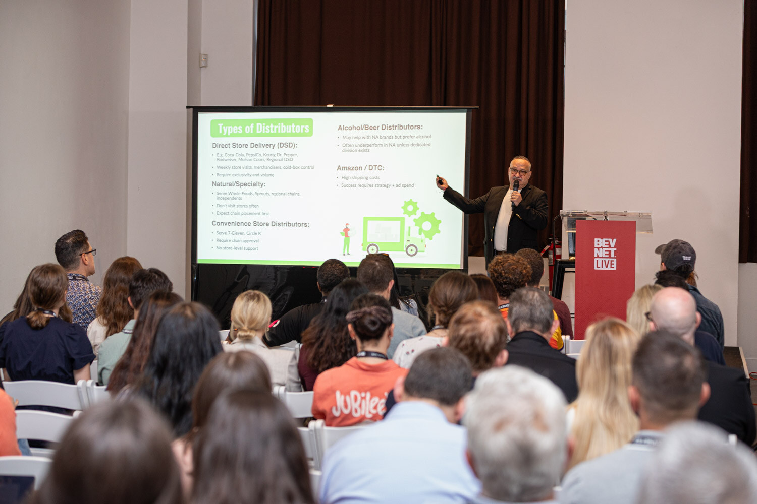 A man points at a green slide titled Types of Distributors while seated attendees watch from the foreground.