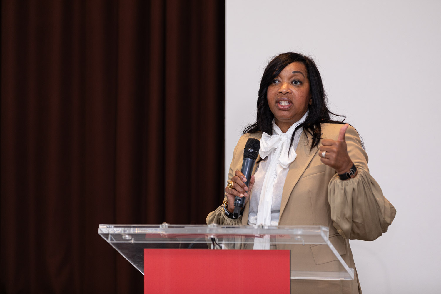 A woman in a tan blazer and white bow blouse speaks into a microphone at a clear podium.