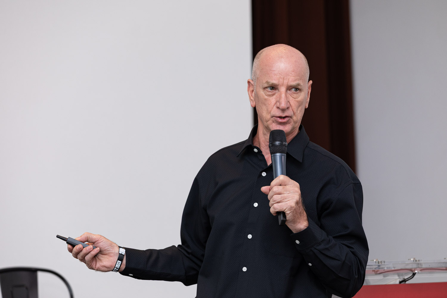 A bald man in a black shirt holds a microphone and slide clicker.
