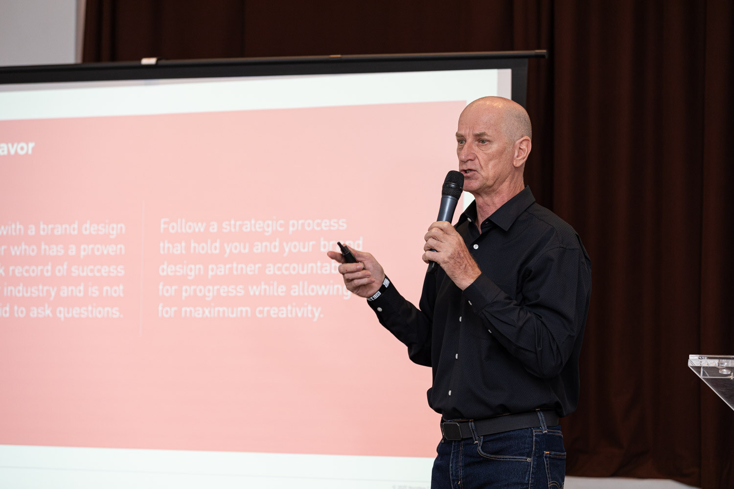 A man in a black shirt holds a microphone beside a pink slide about following a strategic process.