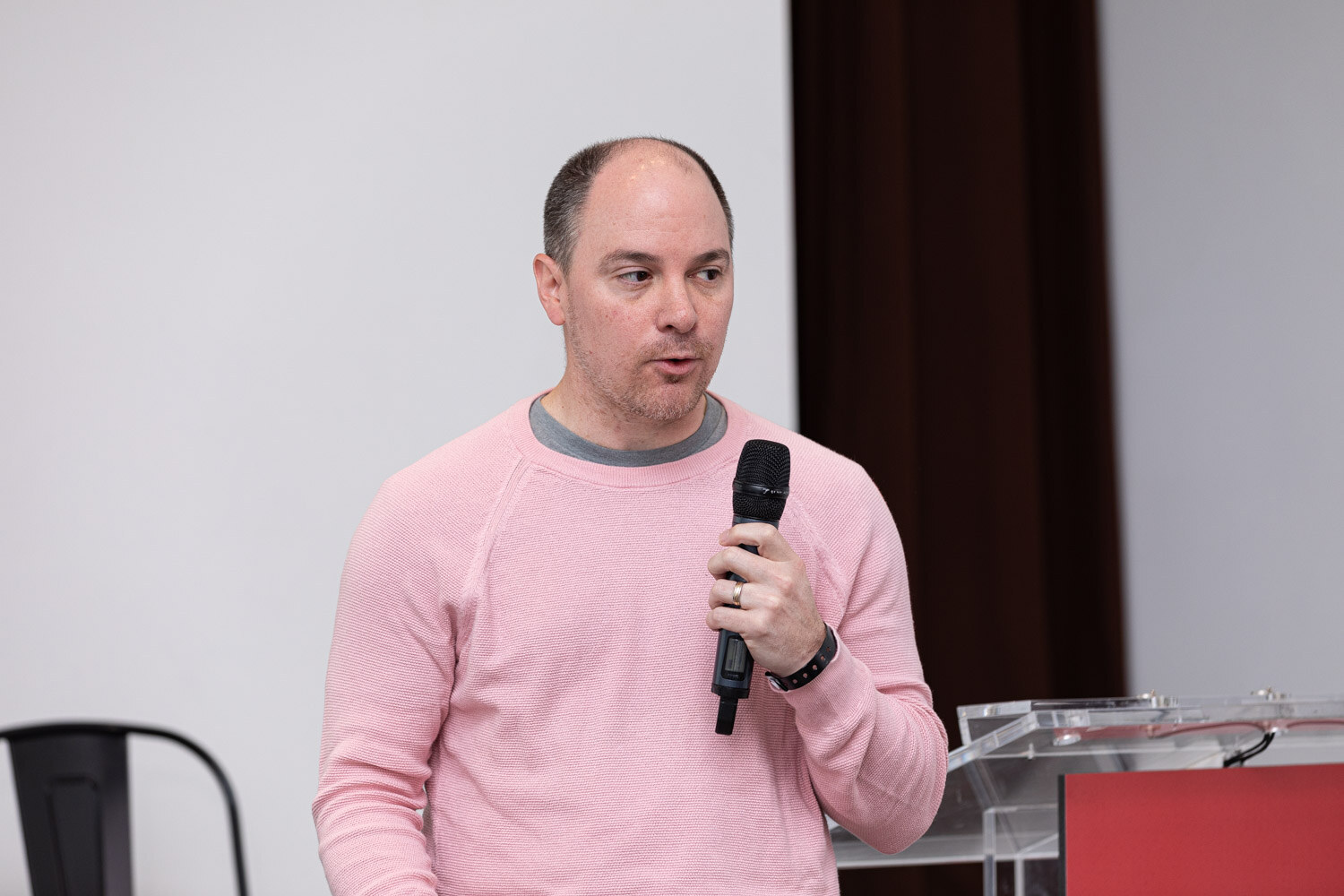 A man in a pink sweater holds a microphone in front of a white wall and dark curtain.