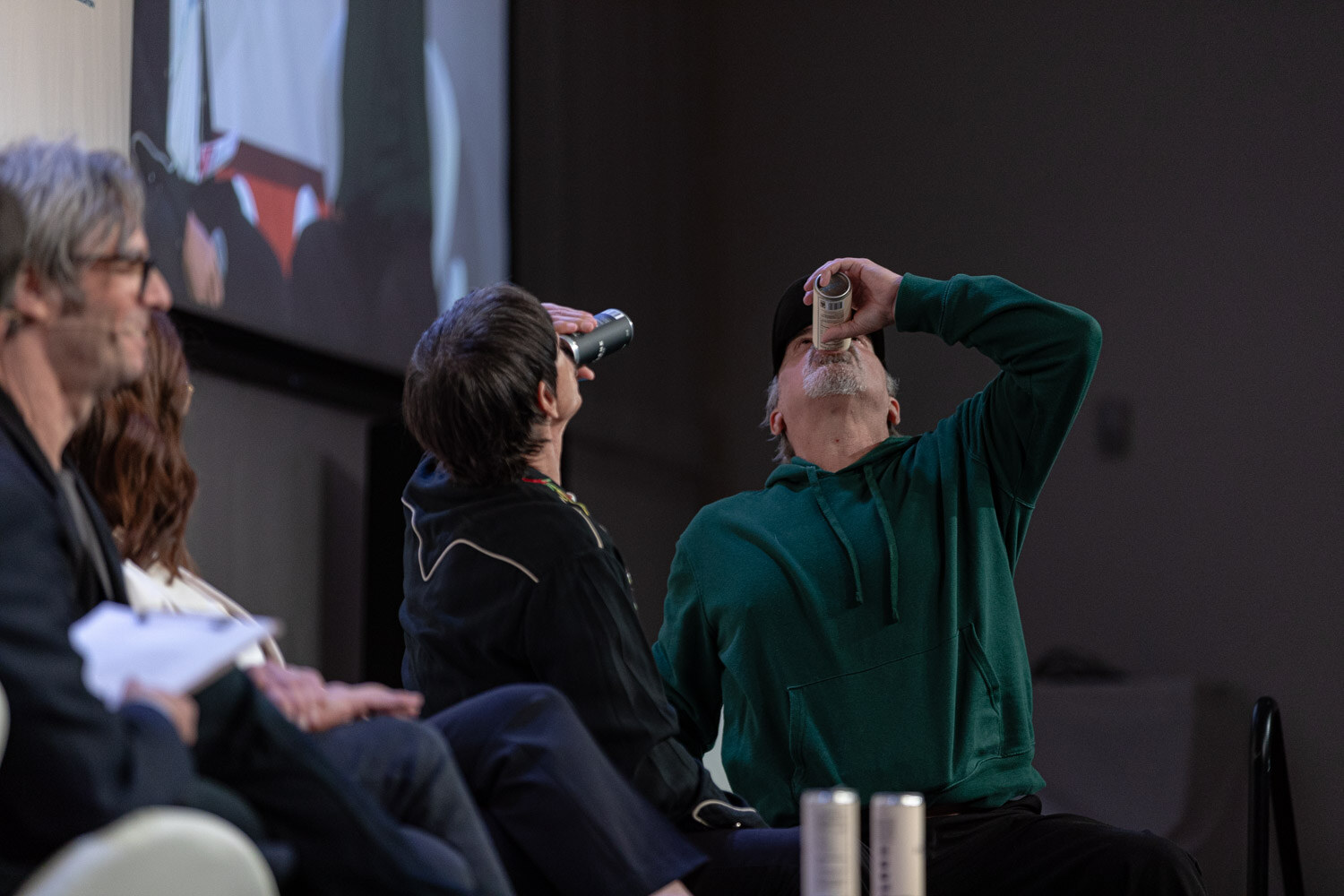 Two seated men on a conference stage drinking from cans, with two people in the left foreground.
