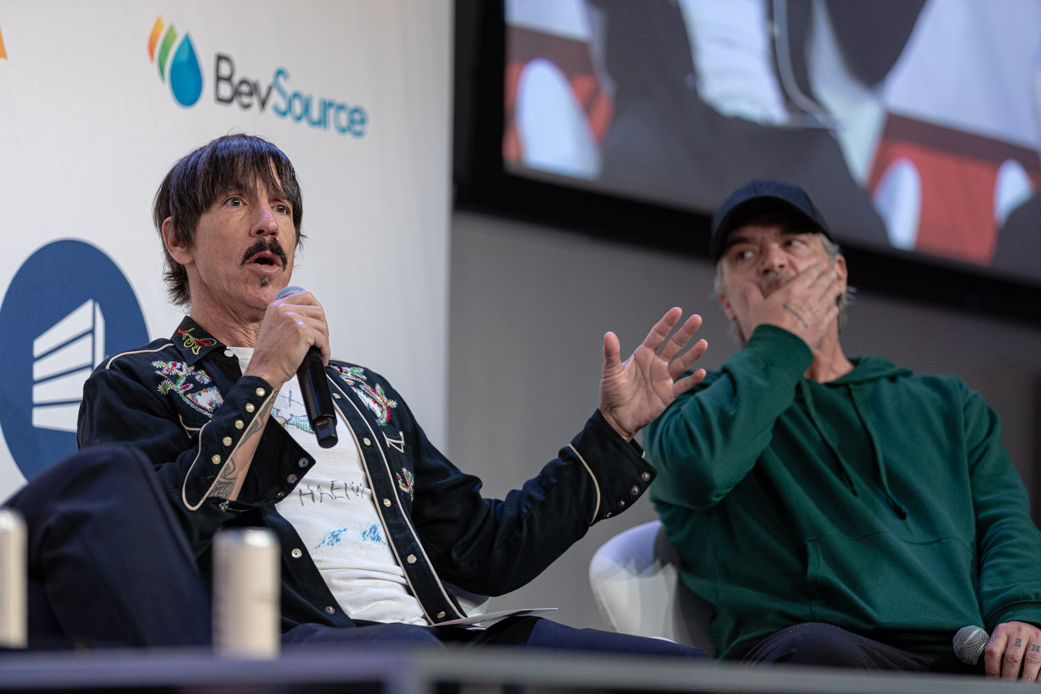 Man with a mustache and dark embroidered jacket holding a microphone, with a man in a cap beside him.