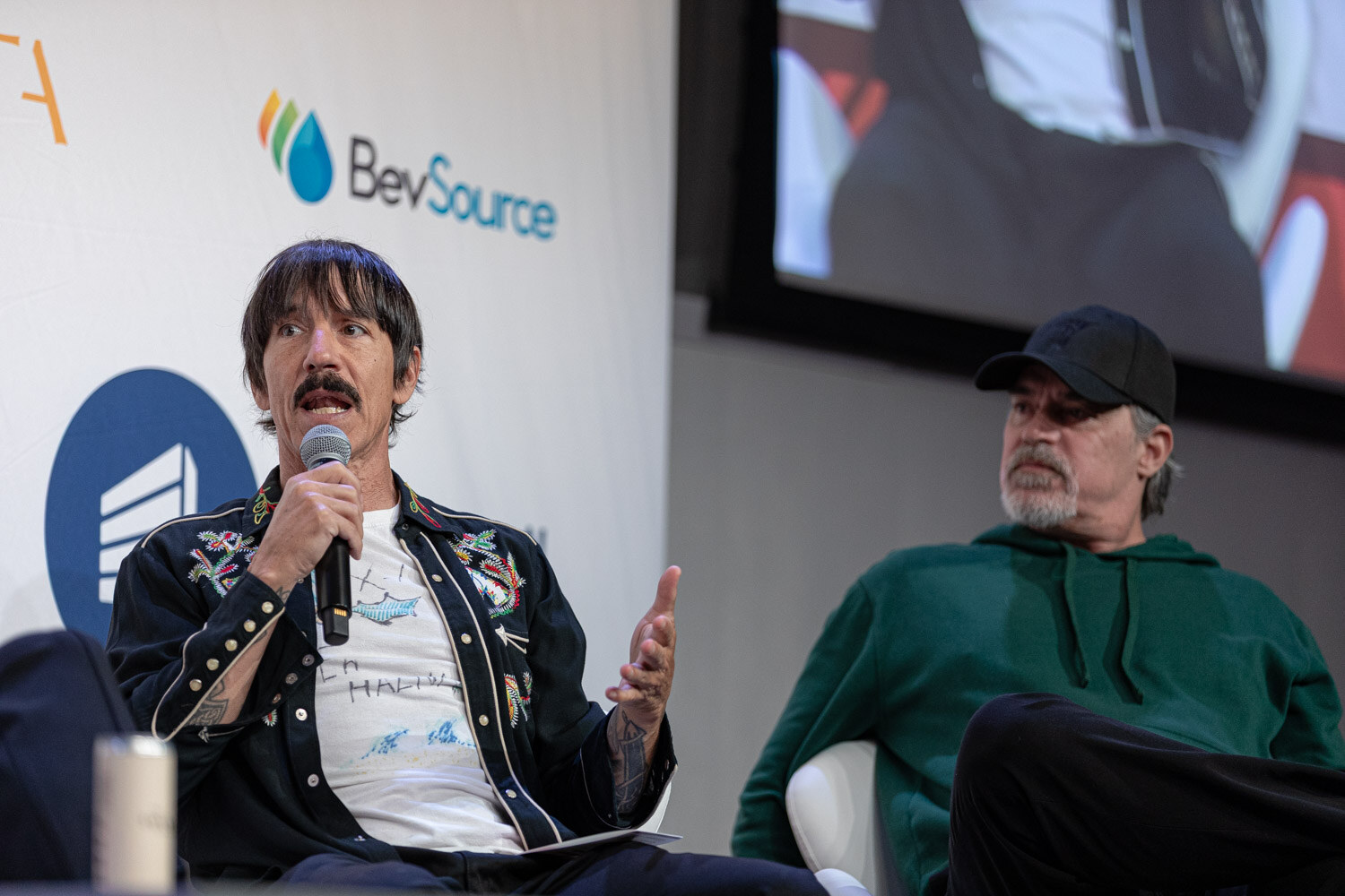 Man in an embroidered jacket speaking into a microphone with a man in a black cap seated behind.