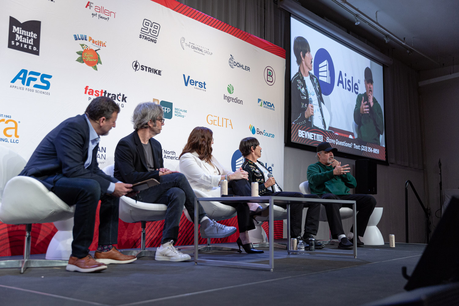 Five people seated on stage in front of a sponsor wall, with a large screen on the right.