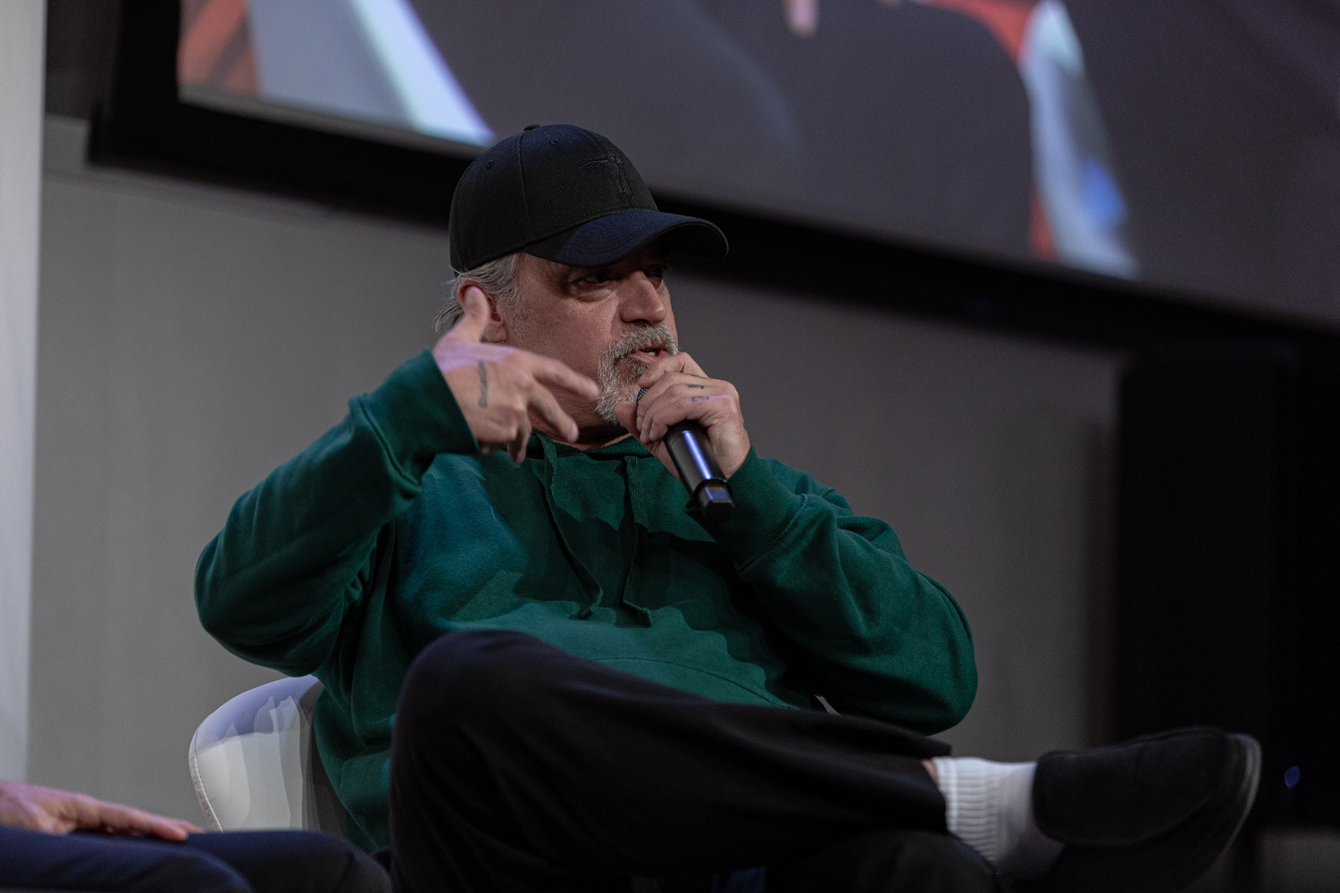 Man in a black cap and green hoodie holding a microphone and gesturing with his hand.
