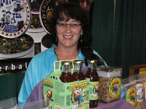 A smiling woman with glasses and dark hair in a light blue shirt holds a green four-pack carton of Jorma's bottled sauce, with tea tins and packaged goods on the table around her.
