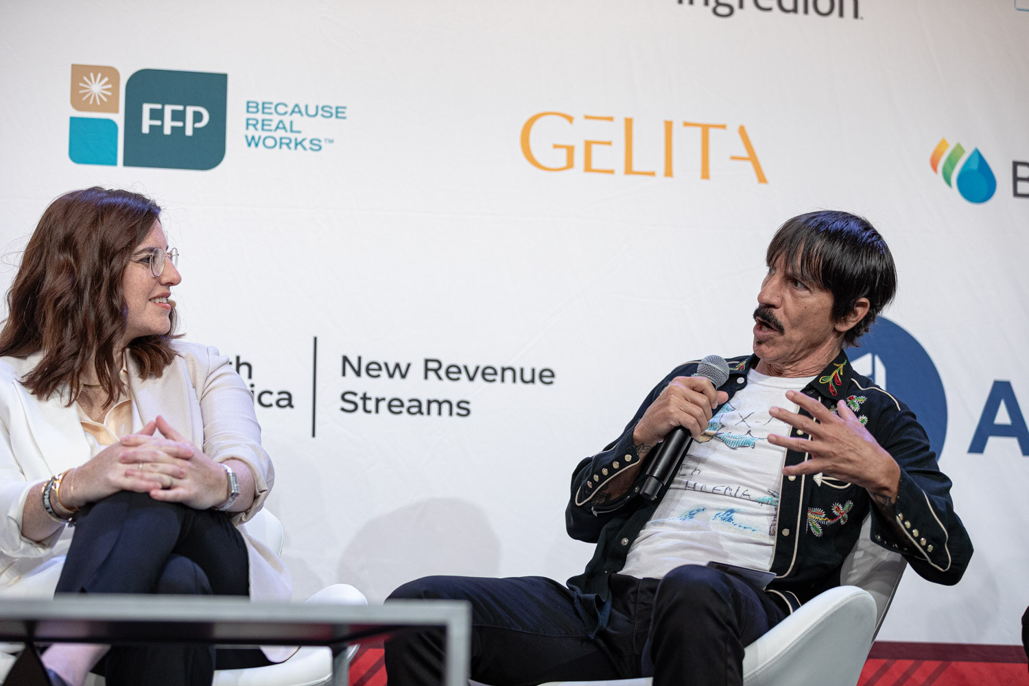 Woman in a white blazer and a man in an embroidered jacket seated in front of sponsor logos and the words New Revenue Streams.