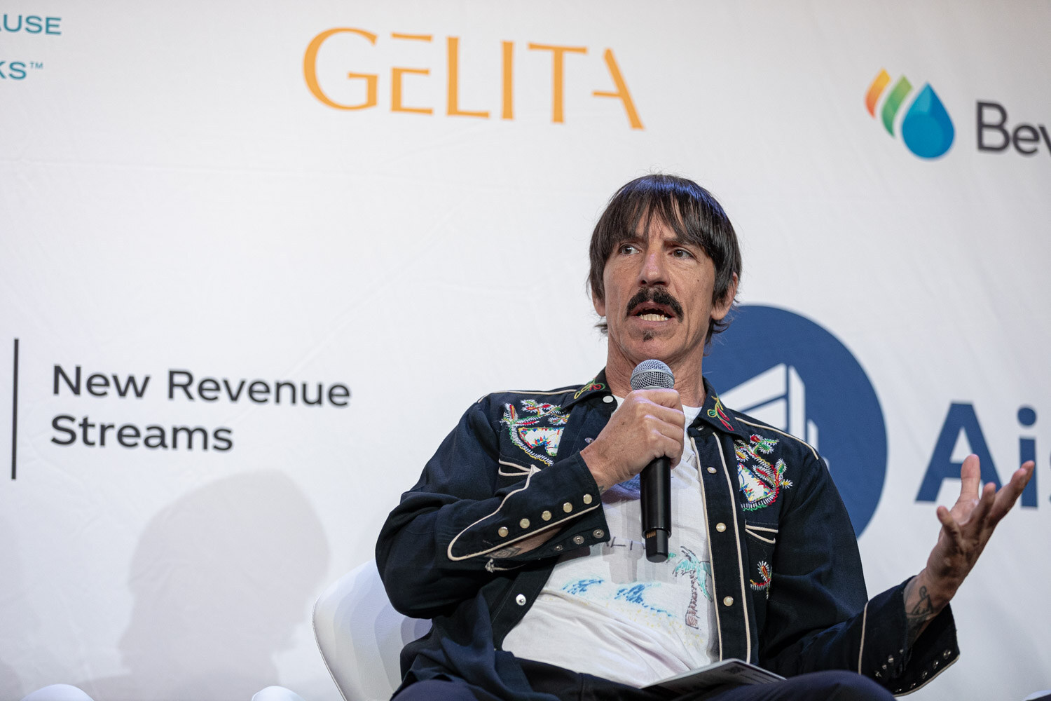 Man in an embroidered jacket holding a microphone in front of a white sponsor wall.