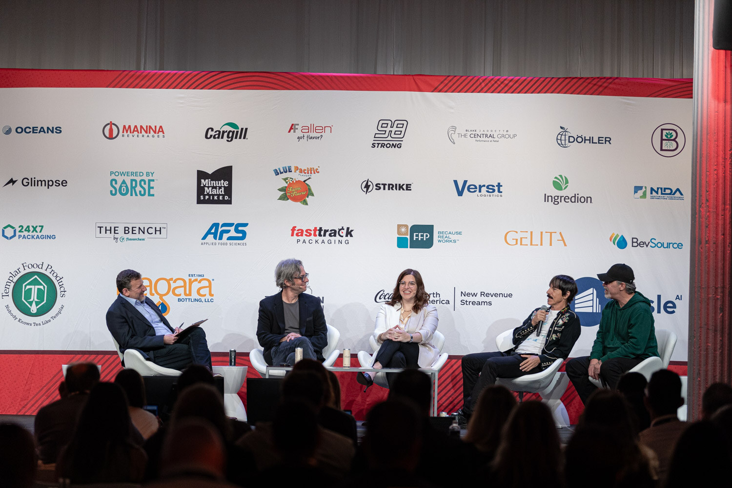 Five people seated in white chairs on stage in front of a sponsor wall with logos including Cargill and Coca-Cola.
