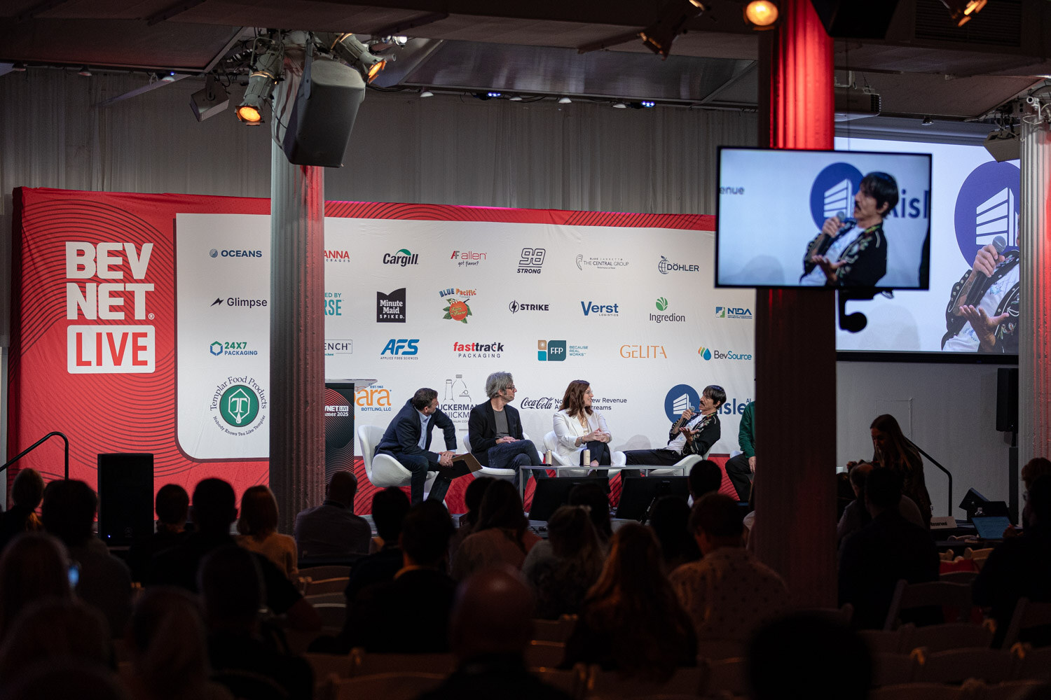 Wide view of a stage with a red BevNET Live sign, sponsor wall and screen showing the panelists.