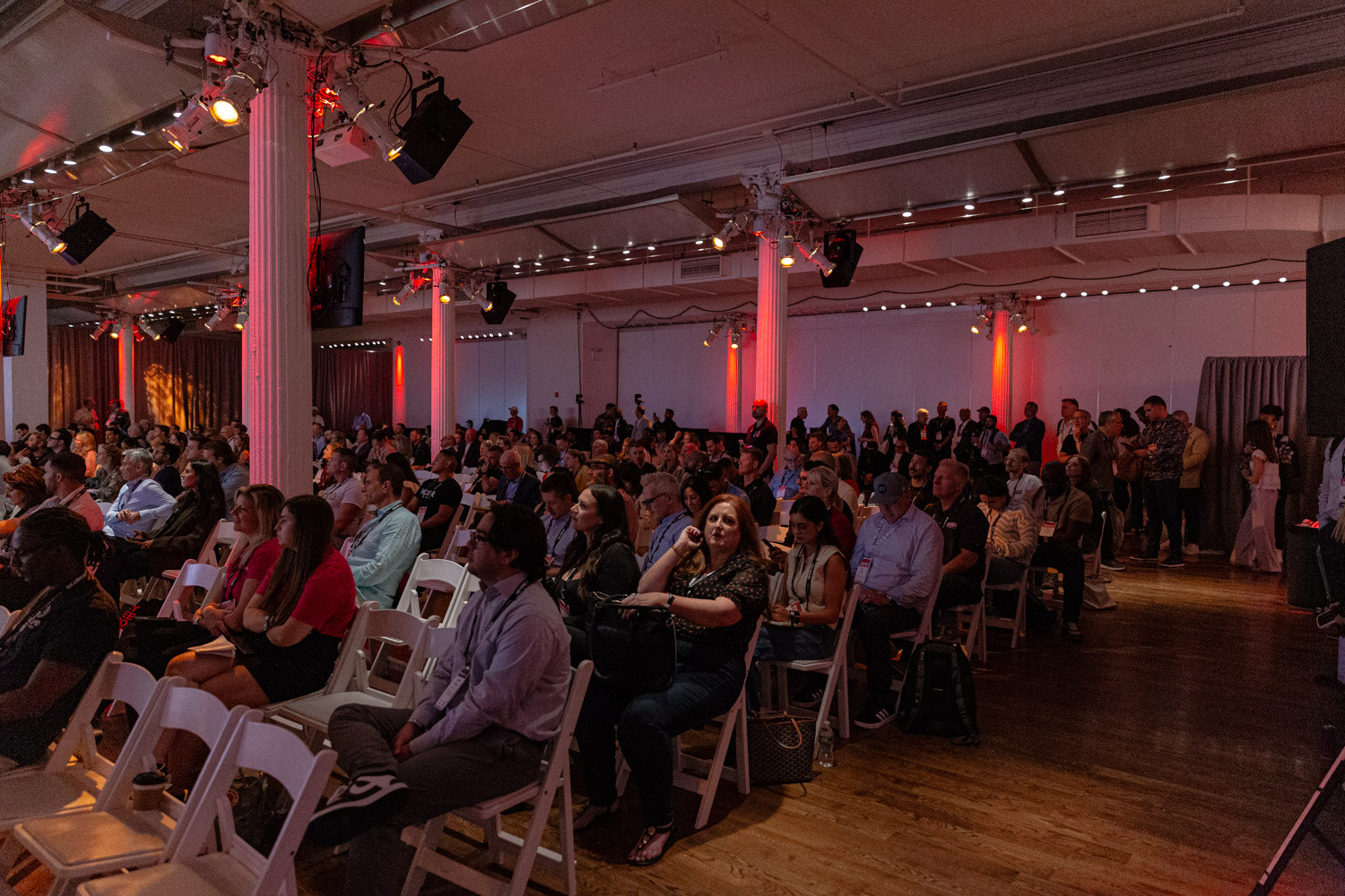 Audience seated in white folding chairs in a ballroom with red uplighting.