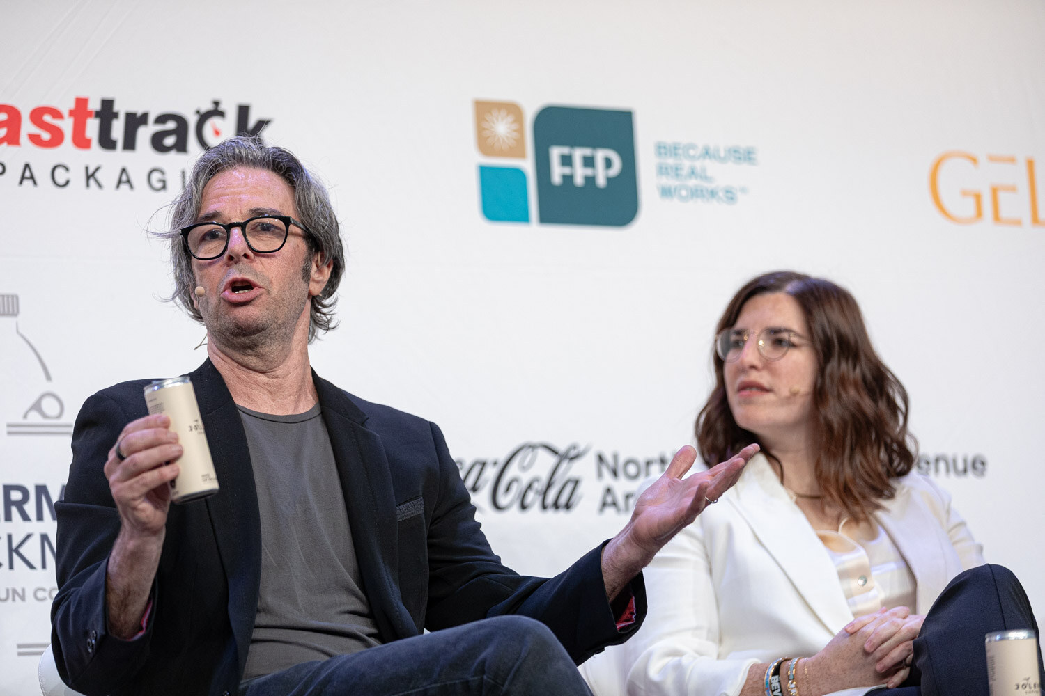 Man in glasses holding a can gesturing while a woman in a white blazer listens.