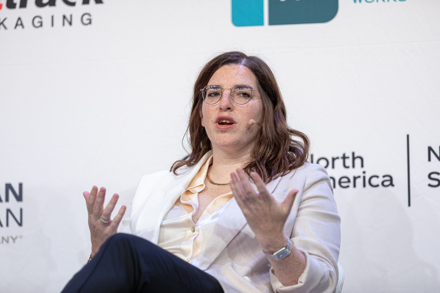 Woman with dark hair and glasses in a cream blouse and white blazer speaking on stage.