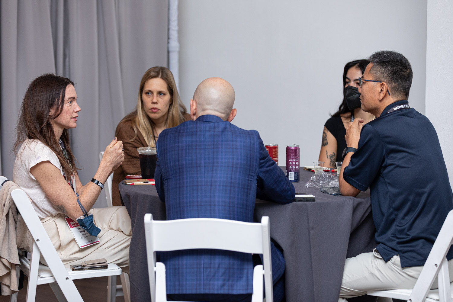 Four people sit around a round table with a gray cloth, with cans on the table, in a room with gray curtains.