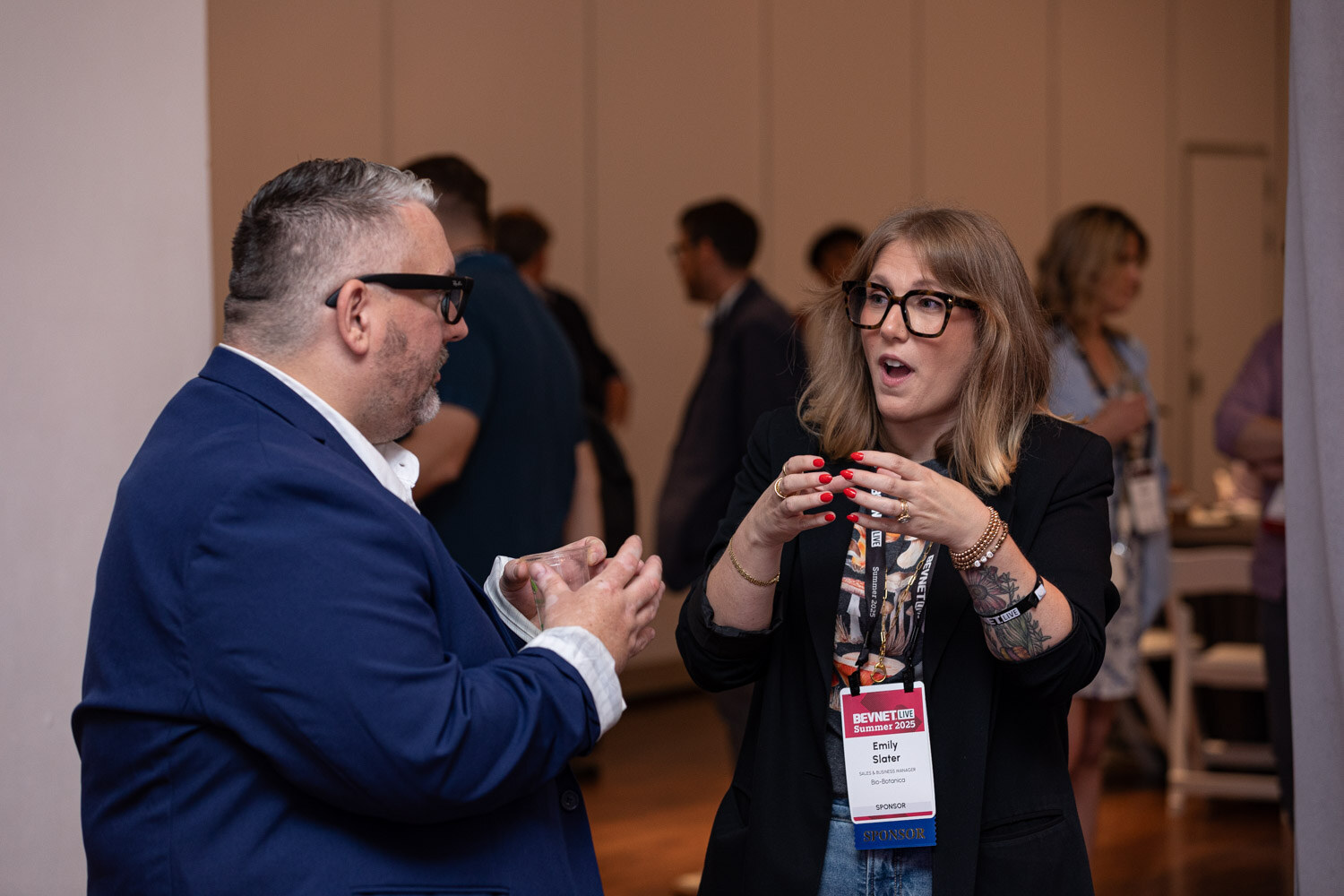 A man in a blue blazer and a woman with glasses and a badge talk face to face in the exhibit hall.