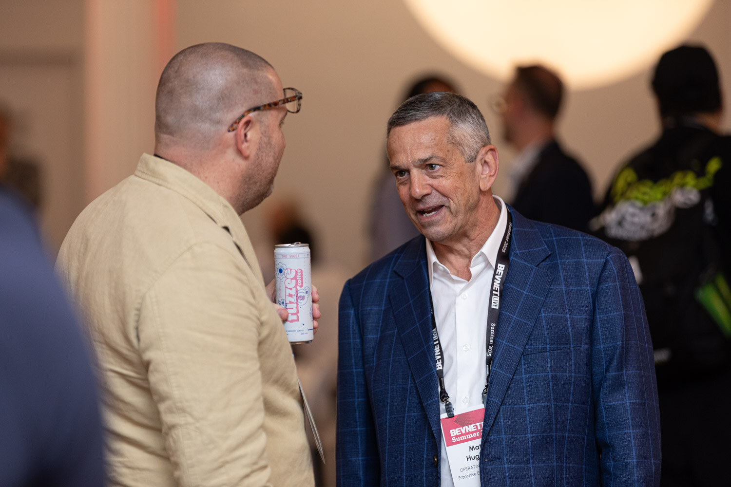 A bald man in a tan shirt holding a pink can talks with a man in a blue plaid blazer wearing a lanyard.