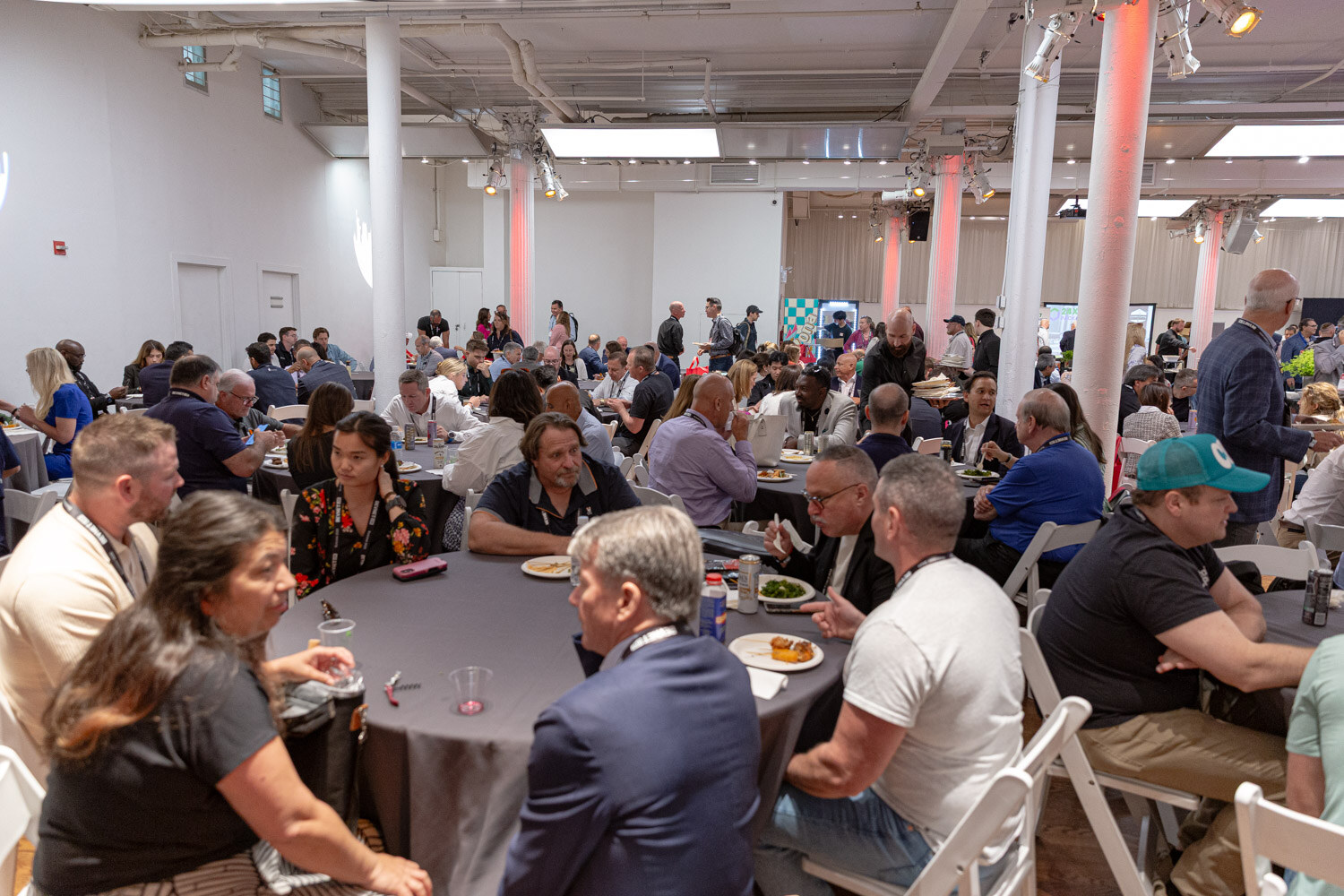 A wide view of a large hall with white columns and round tables full of attendees eating lunch.