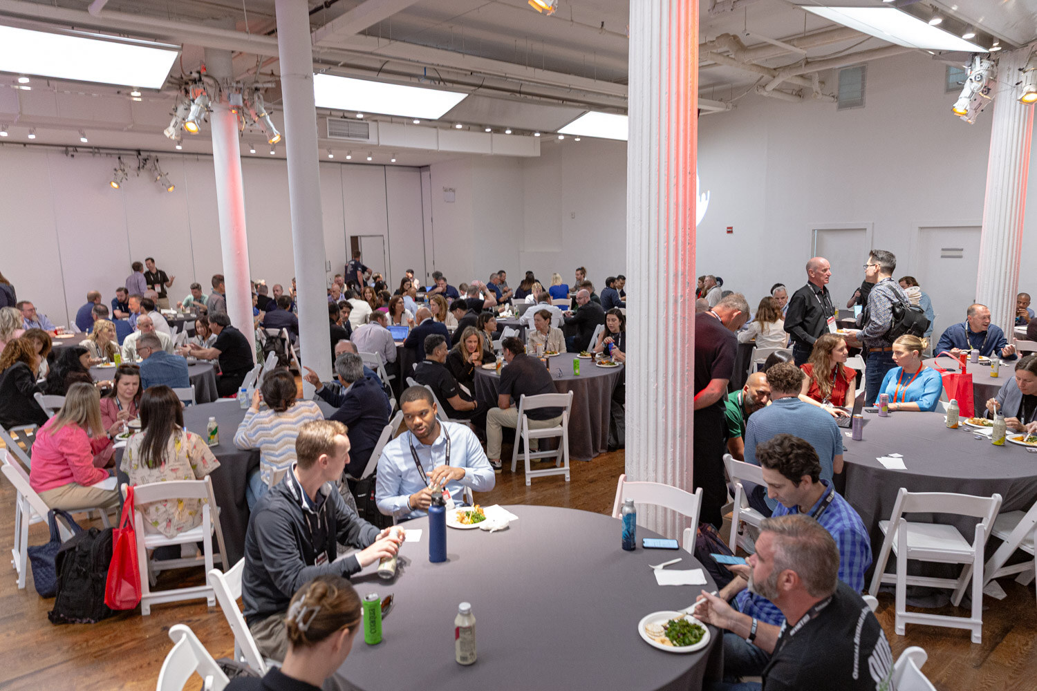 A wide view of a hall with white columns, round tables and seated attendees with lanyards.