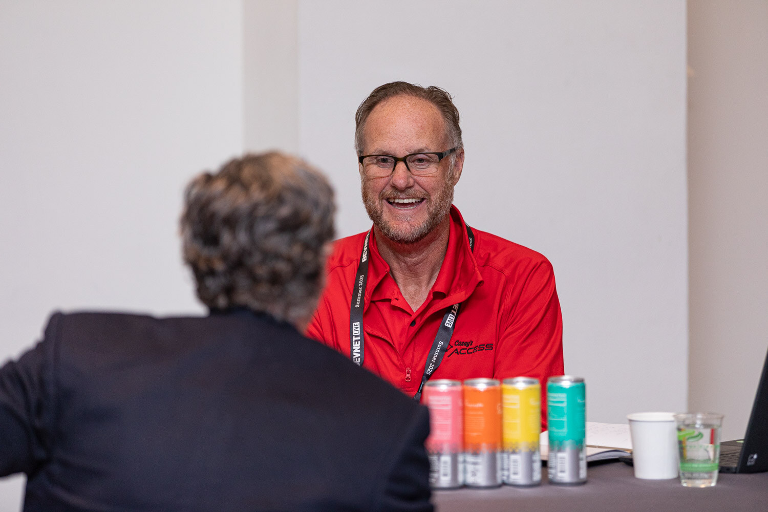 A man in a red polo and glasses smiles at a table with four colorful cans, across from a man with his back to the camera.