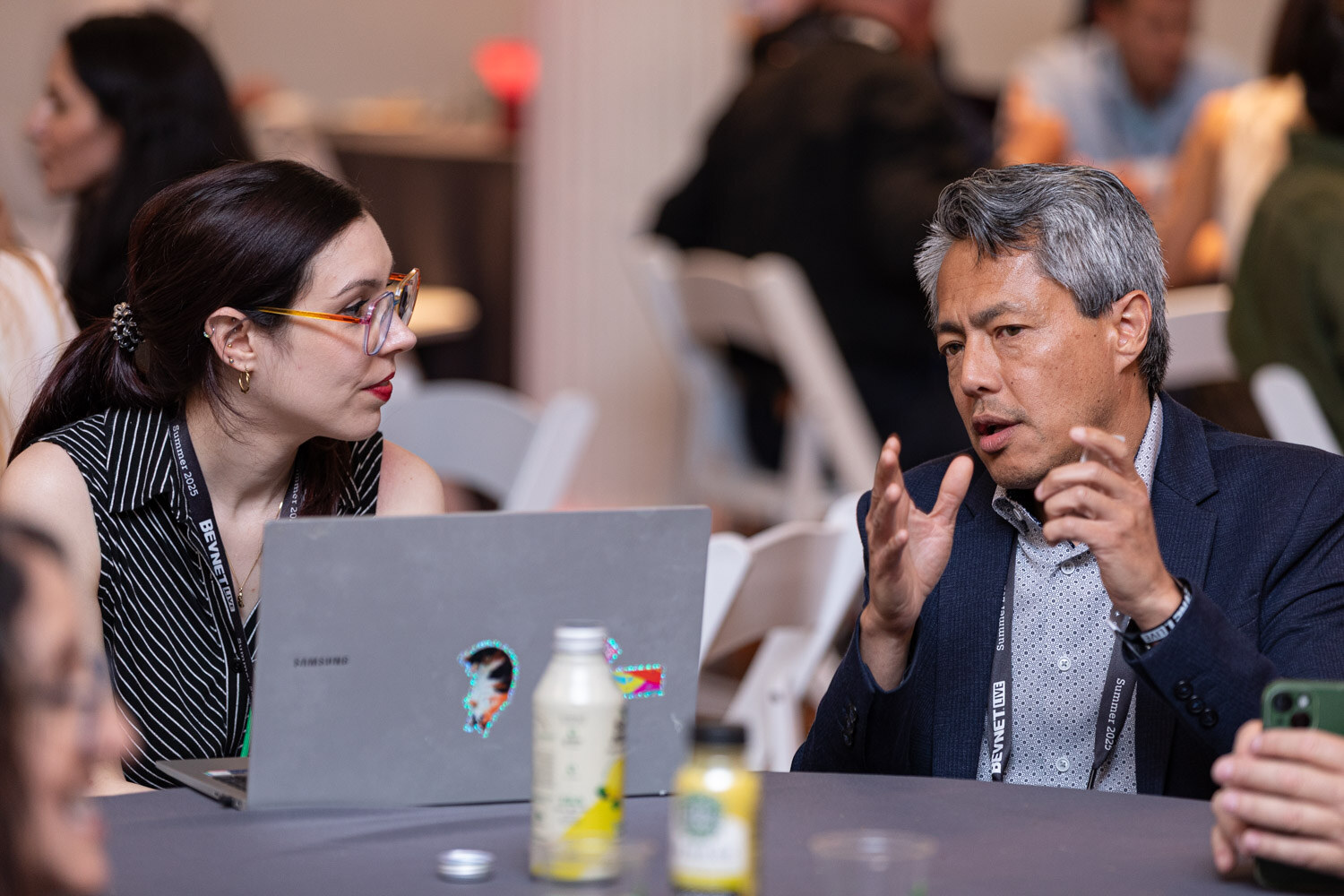 A woman with glasses and a man with gray hair in a blazer talk at a table with a laptop and two bottled drinks.