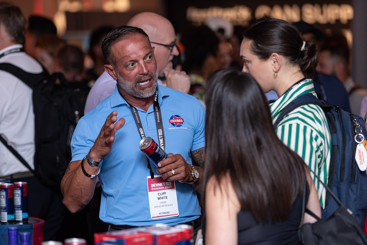 A man in a light blue polo gestures while talking to two women in a crowded hall.