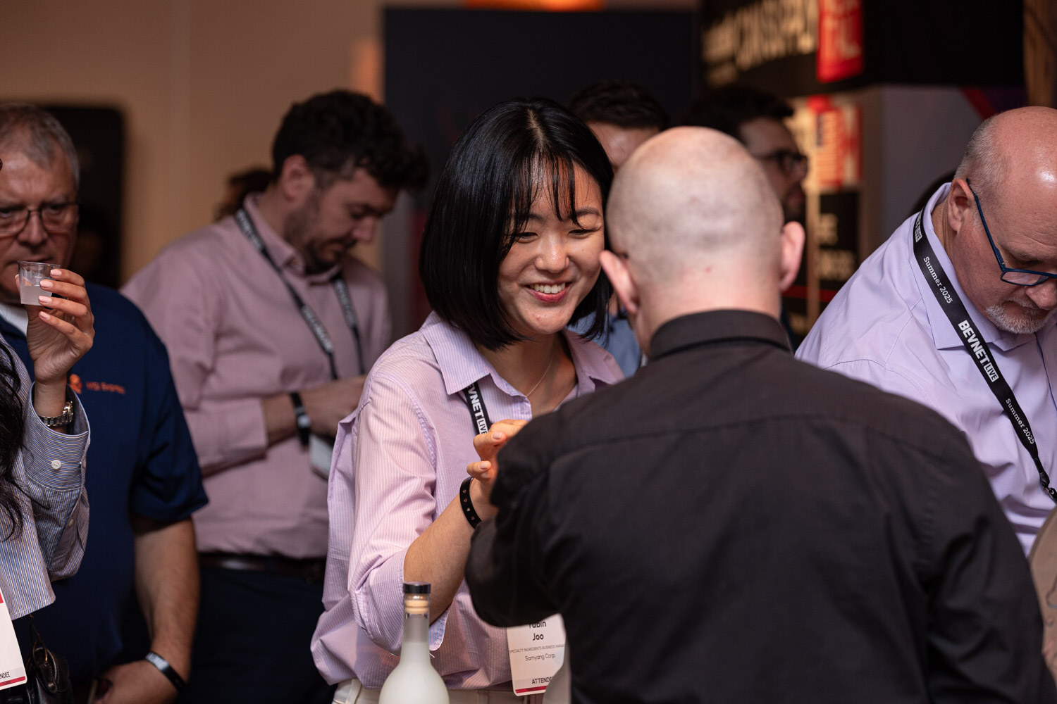 A woman in a pink shirt smiles at a bald man in black while others hold sample cups behind her.