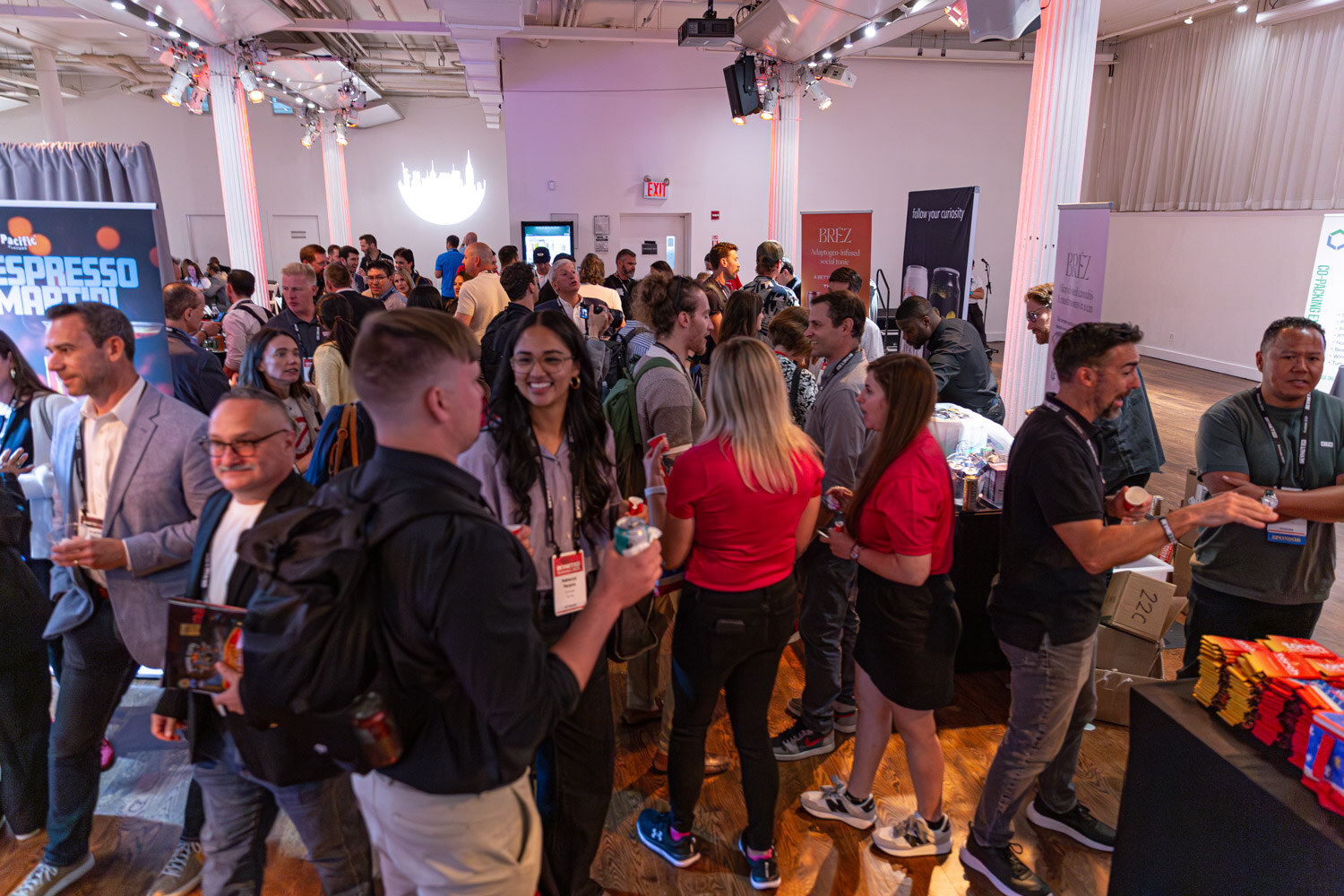 A crowd of attendees with lanyards talk in a hall with exhibitor tables and colorful booths.