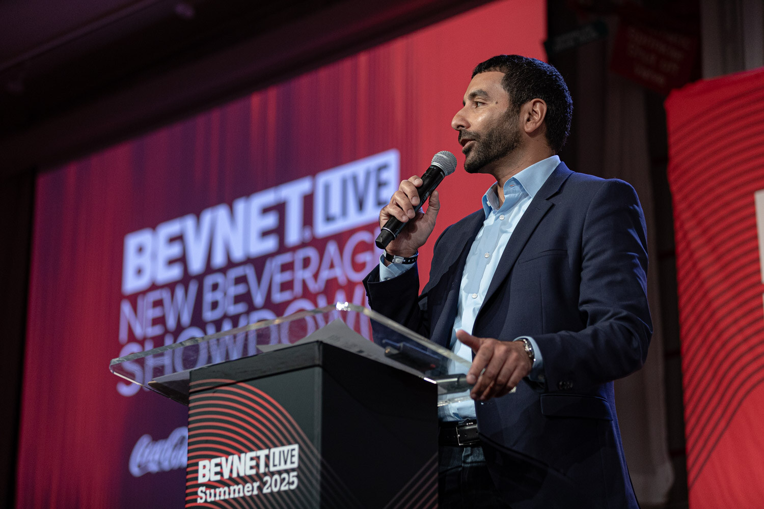 A man in a navy blazer speaks into a handheld microphone at a podium marked BevNET Live Summer 2025.