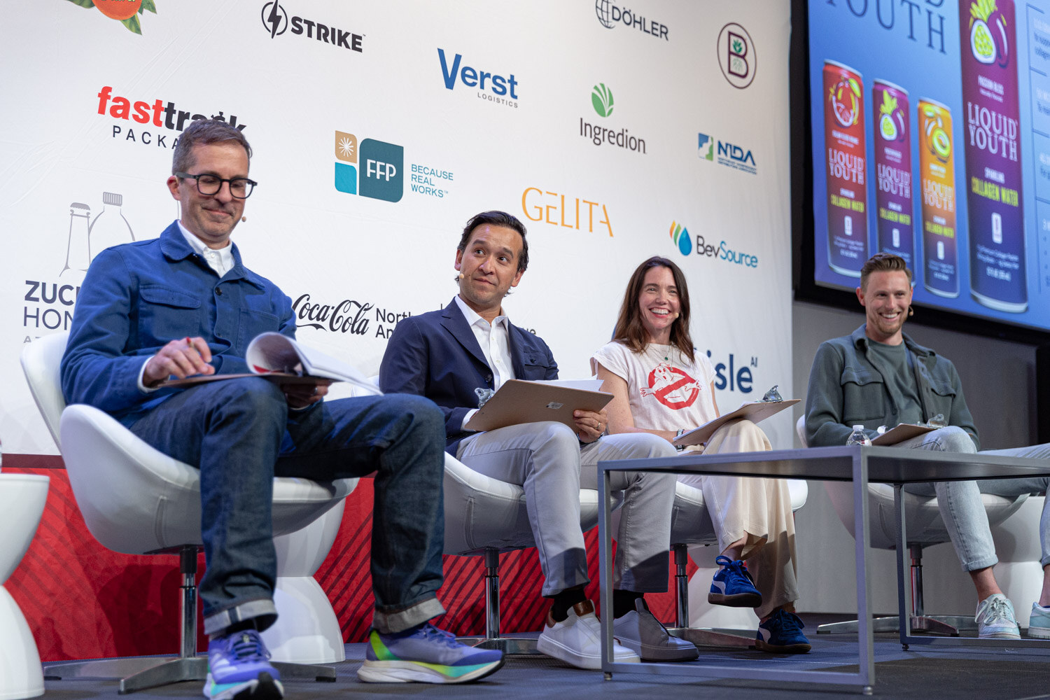 Four people in white chairs on a stage with clipboards, in front of a sponsor backdrop and a Liquid Youth slide.