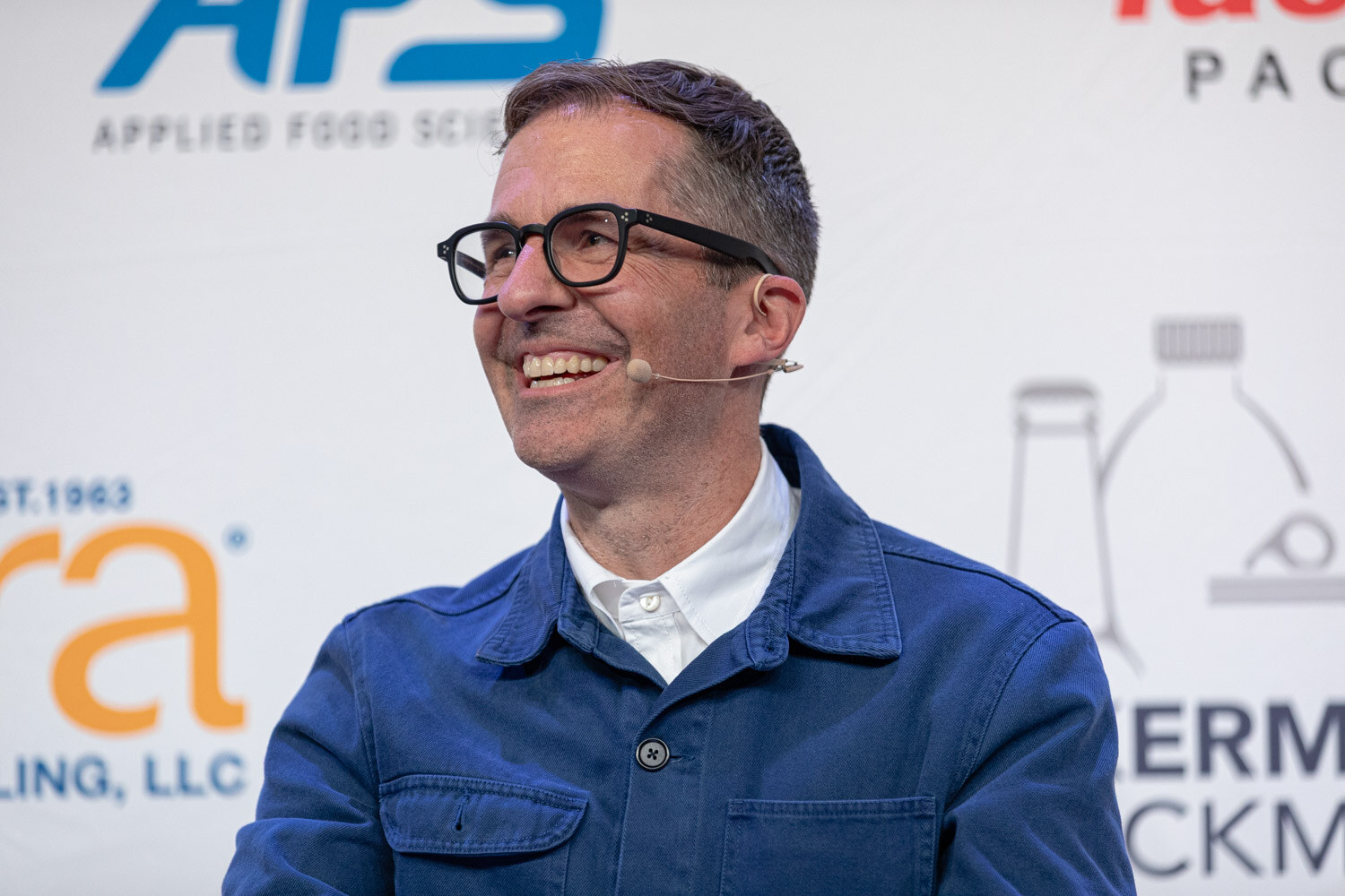 A man with dark glasses and a blue jacket smiles broadly in front of a white sponsor backdrop.