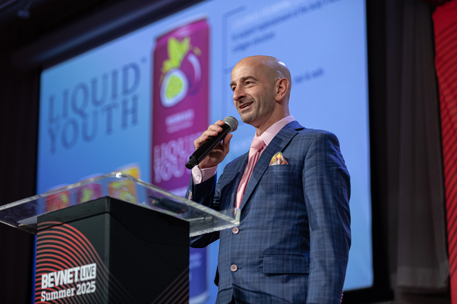 A man in a blue plaid blazer and pink tie holds a microphone at a podium in front of a slide showing Liquid Youth cans.
