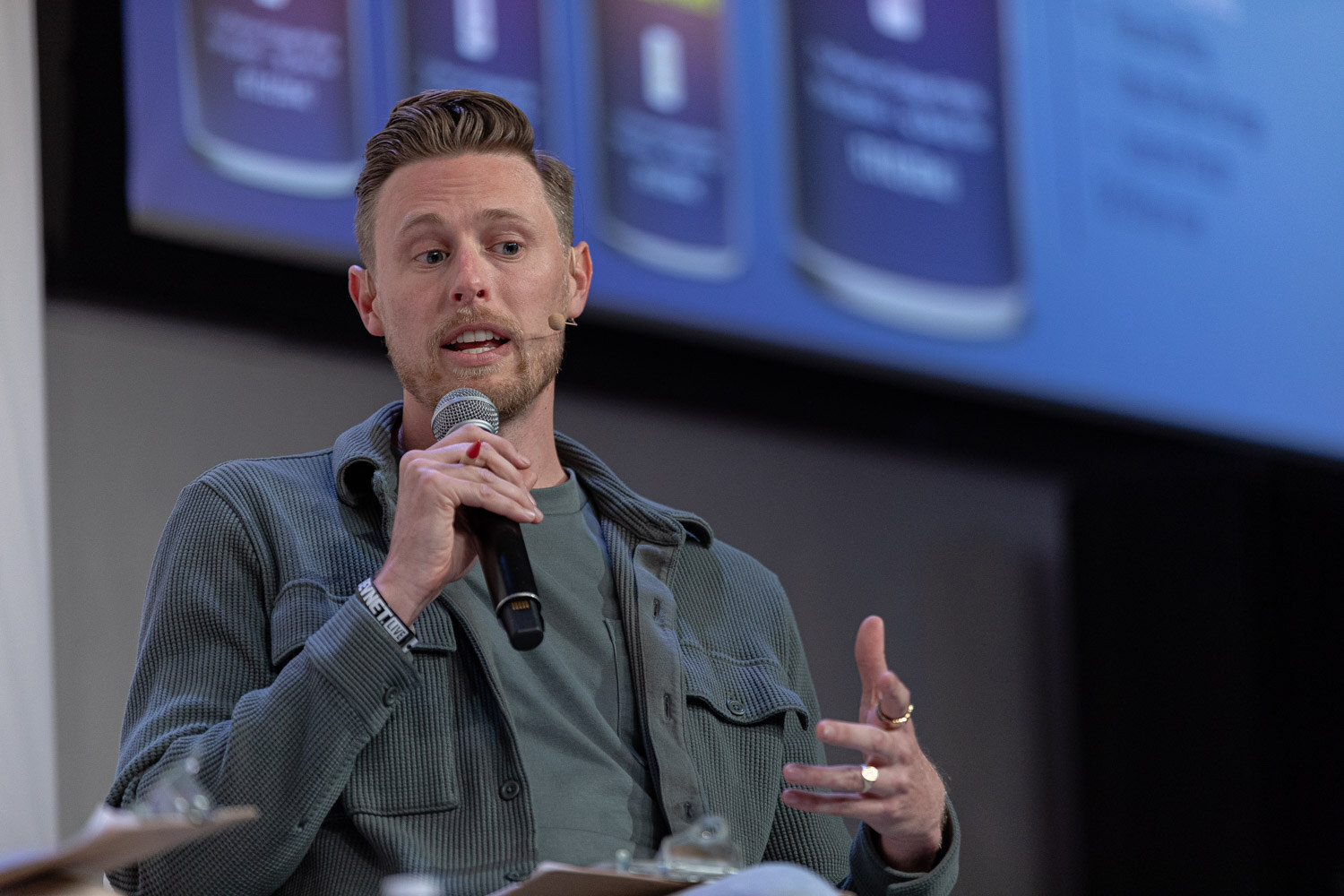 A man in a green-gray overshirt holds a microphone and gestures in front of a screen with product images.