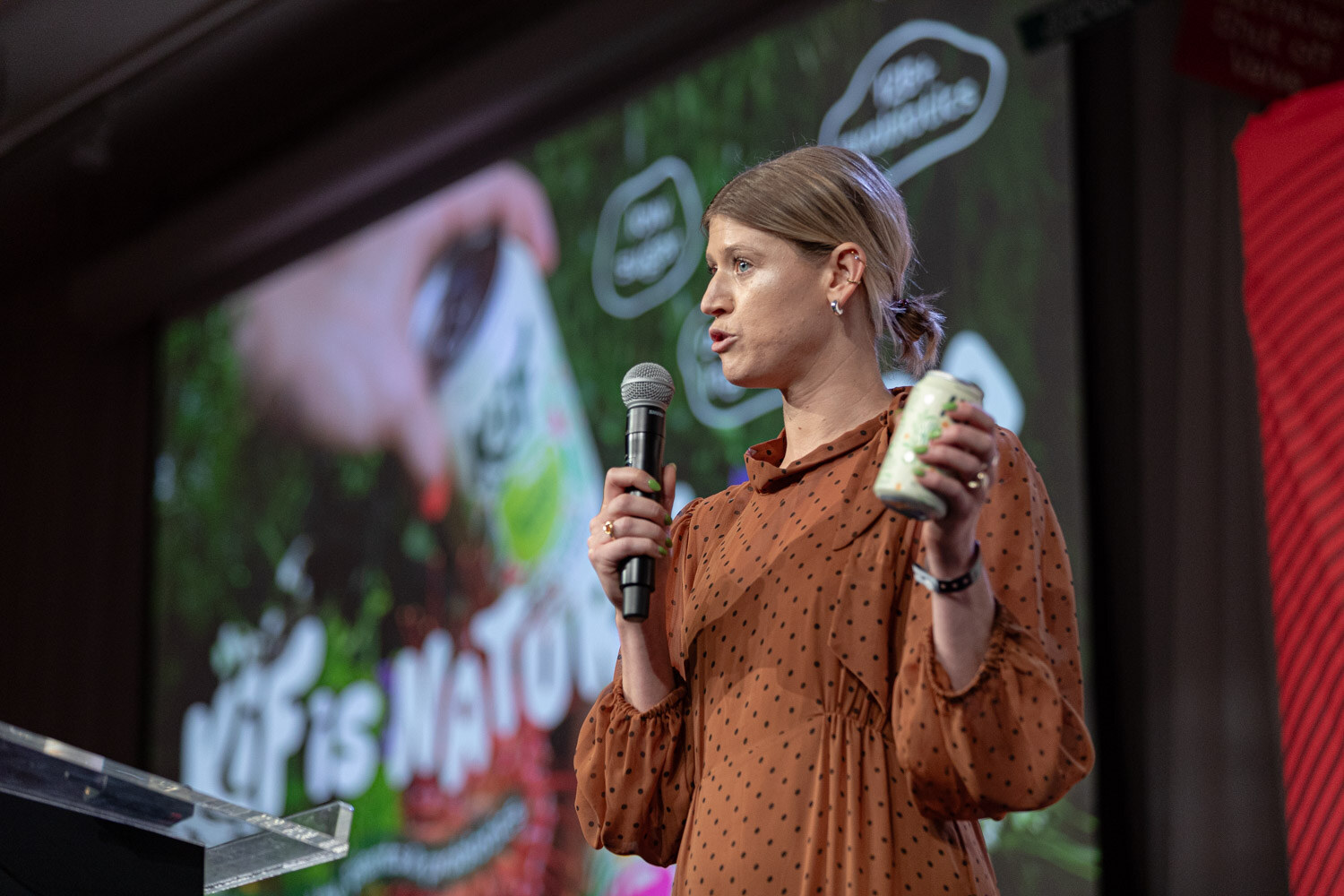 A woman in a rust-colored dress holds a microphone and a can in front of a screen reading Kif is Natural.