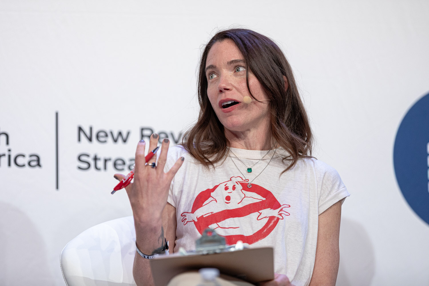 A woman in a Ghostbusters logo t-shirt holds a red pen and a clipboard while looking off to the side.