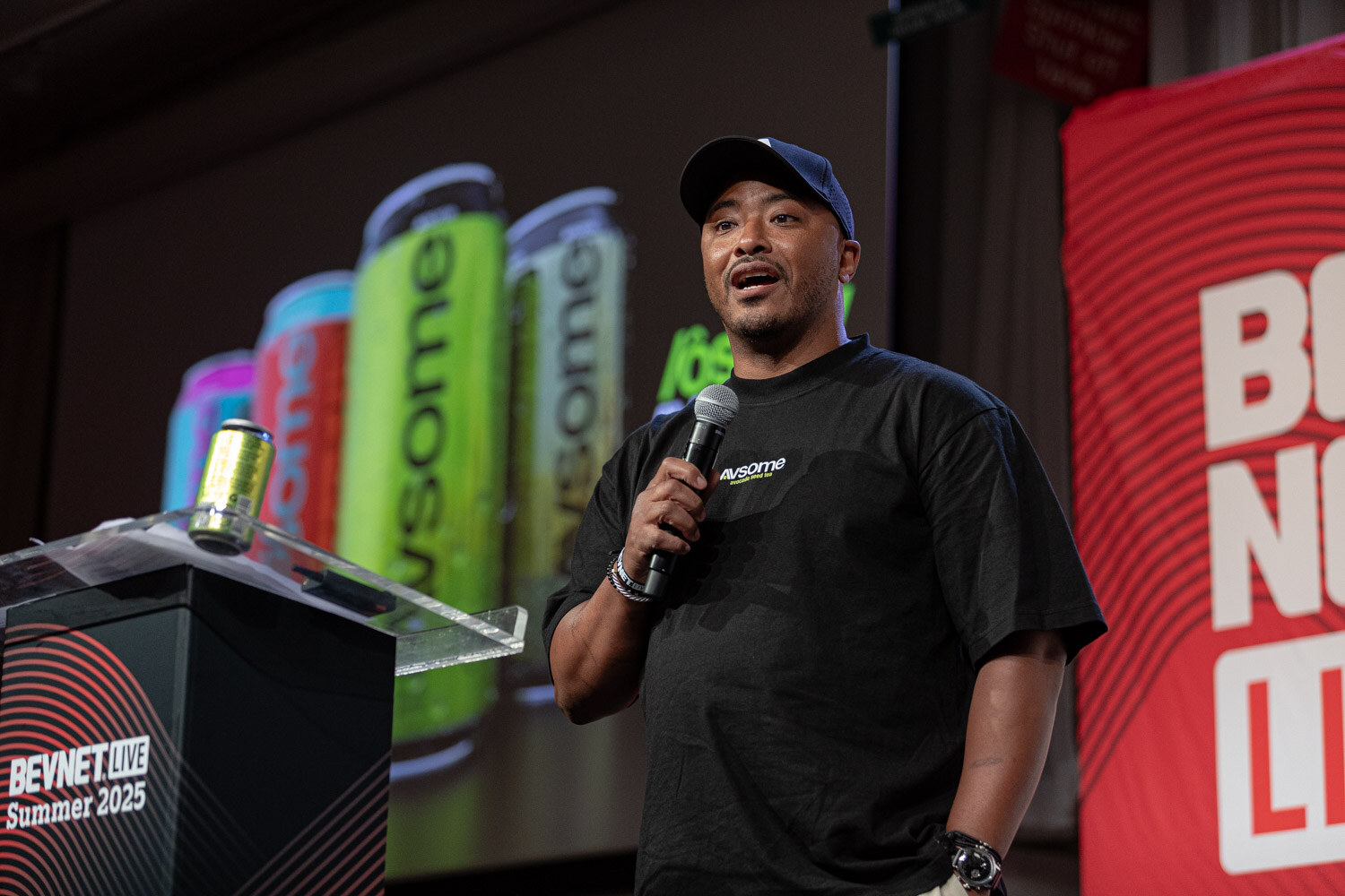 A man in a black AVSOME t-shirt speaks into a microphone beside a podium with a can on it and AVSOME cans on the screen.