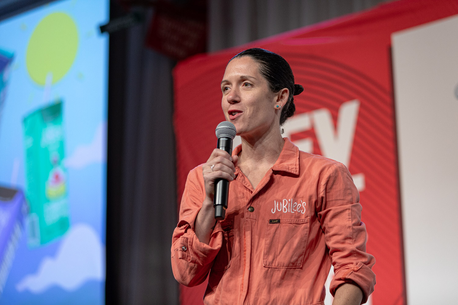 A woman in an orange jumpsuit with Jubilee's on the chest speaks into a microphone in front of a colorful slide.