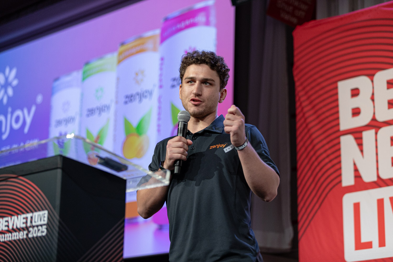 A man in a dark polo gestures while holding a microphone in front of a screen showing Zenjoy cans.