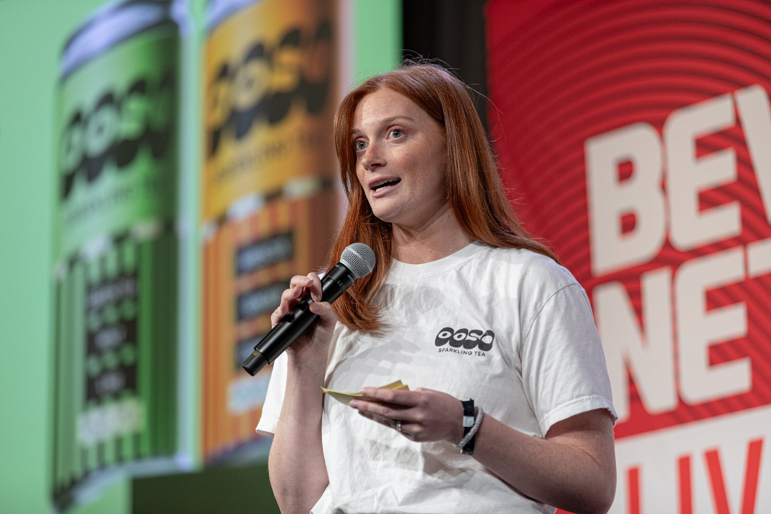 A woman with long red hair and a white OOSO t-shirt holds a microphone in front of a screen showing green and orange OOSO cans.