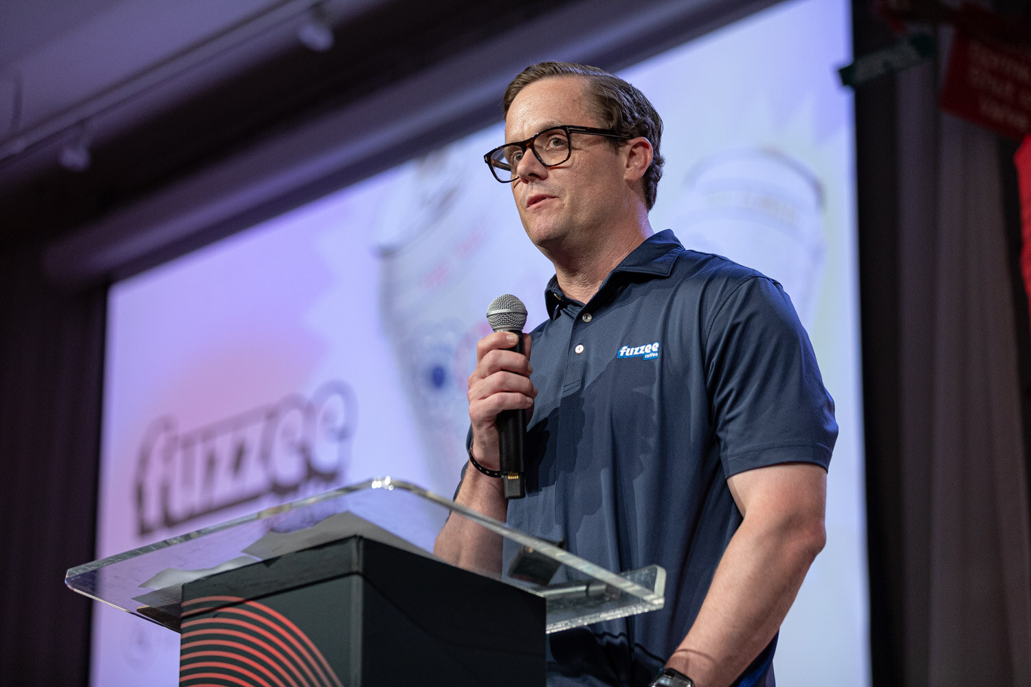 A man in glasses and a navy polo speaks into a microphone in front of a screen showing the Fuzzee logo.