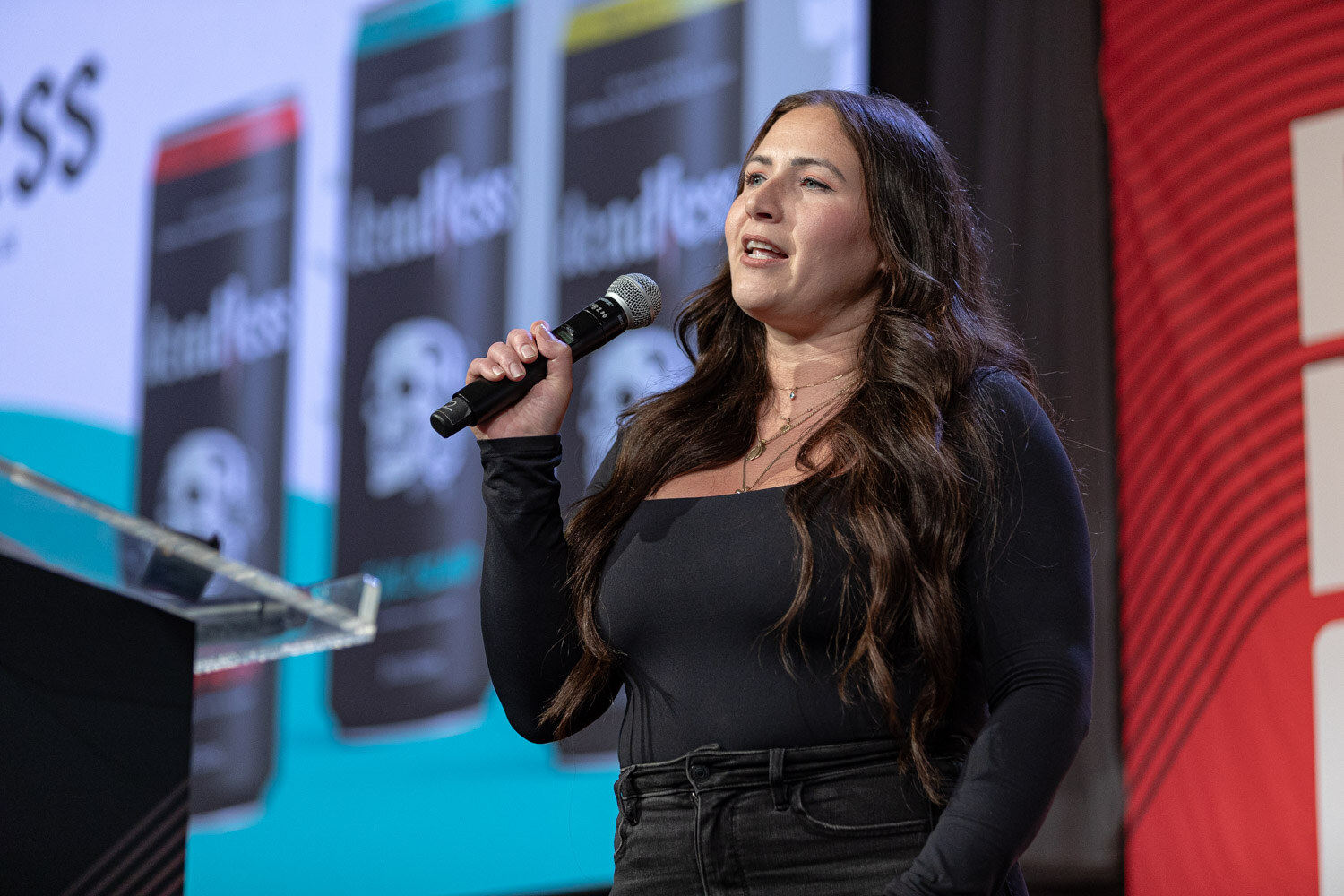 A woman in a black top with long dark hair speaks into a microphone in front of a slide showing black Deadless cans.