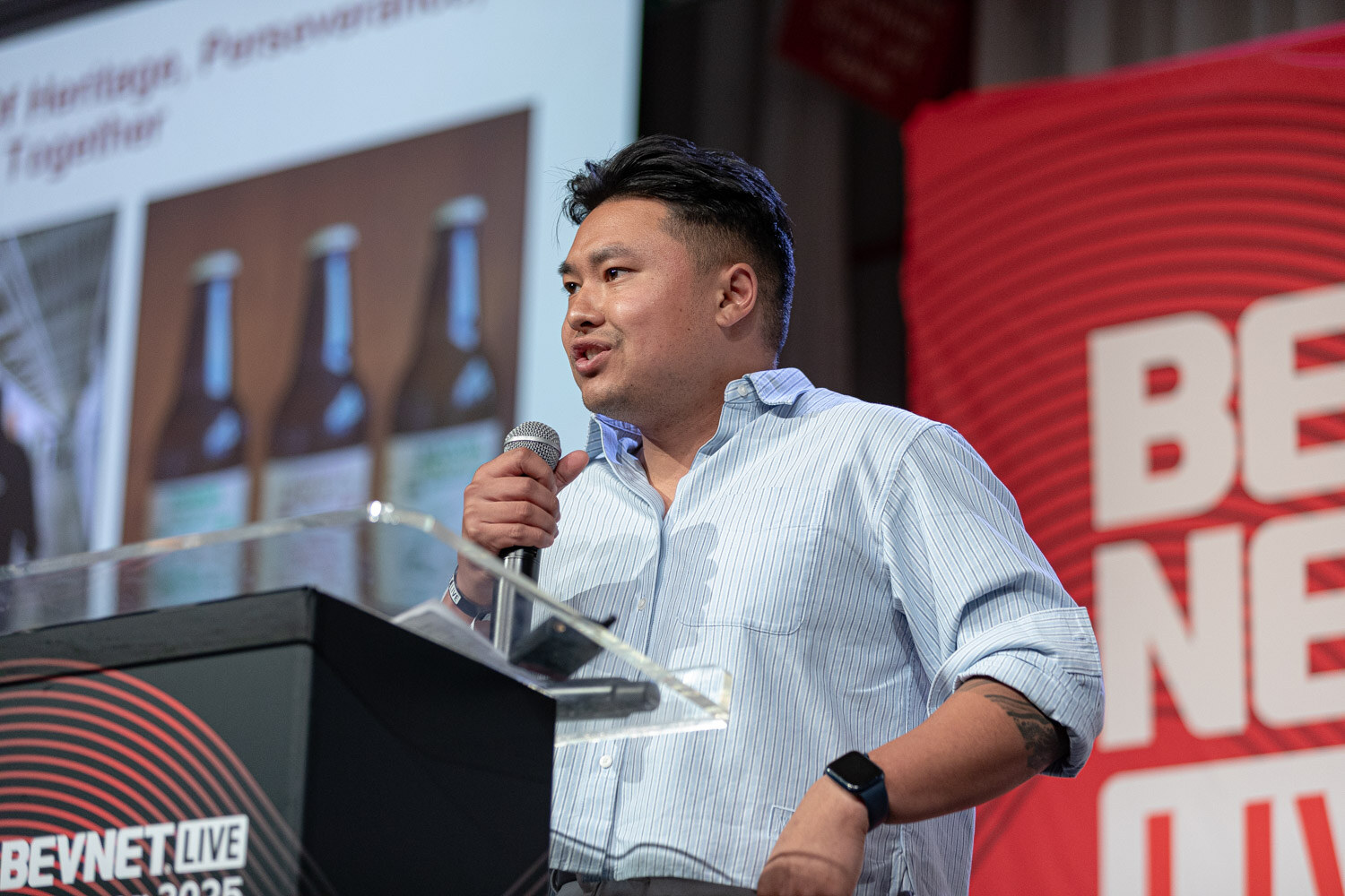 A man in a light blue striped shirt holds a microphone at the podium in front of a slide showing three brown bottles.