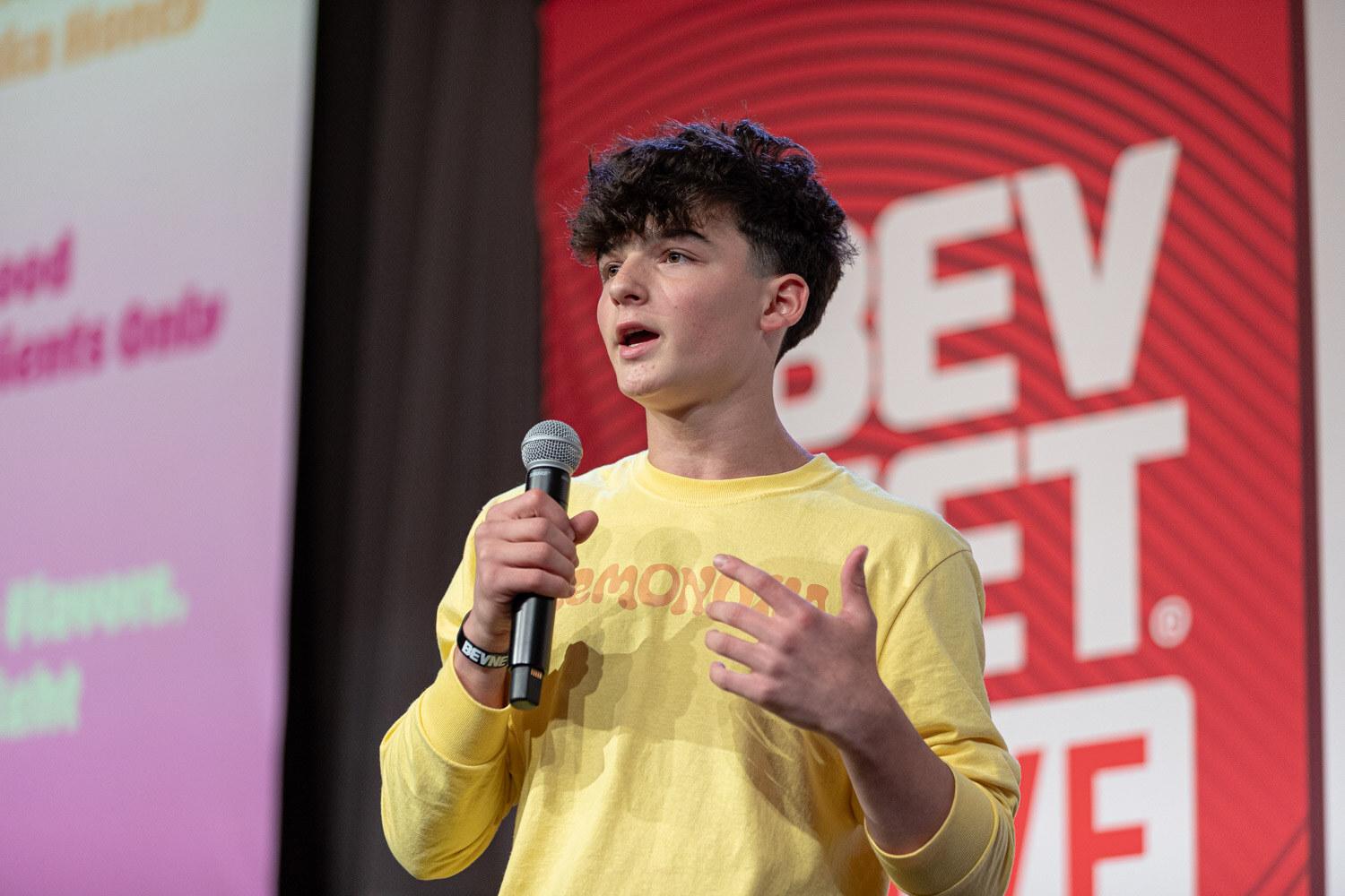 A young man with curly dark hair in a yellow sweatshirt speaks into a microphone in front of a pastel slide.