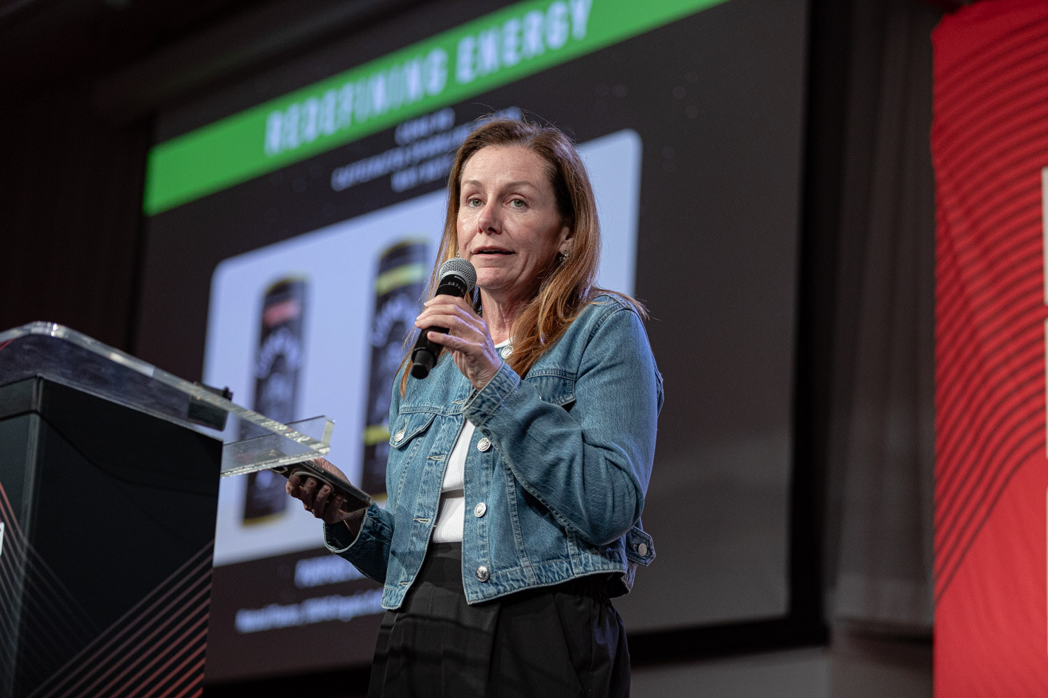 A woman in a denim jacket holds a microphone and phone beside a podium, with a green Redefining Energy slide behind her.
