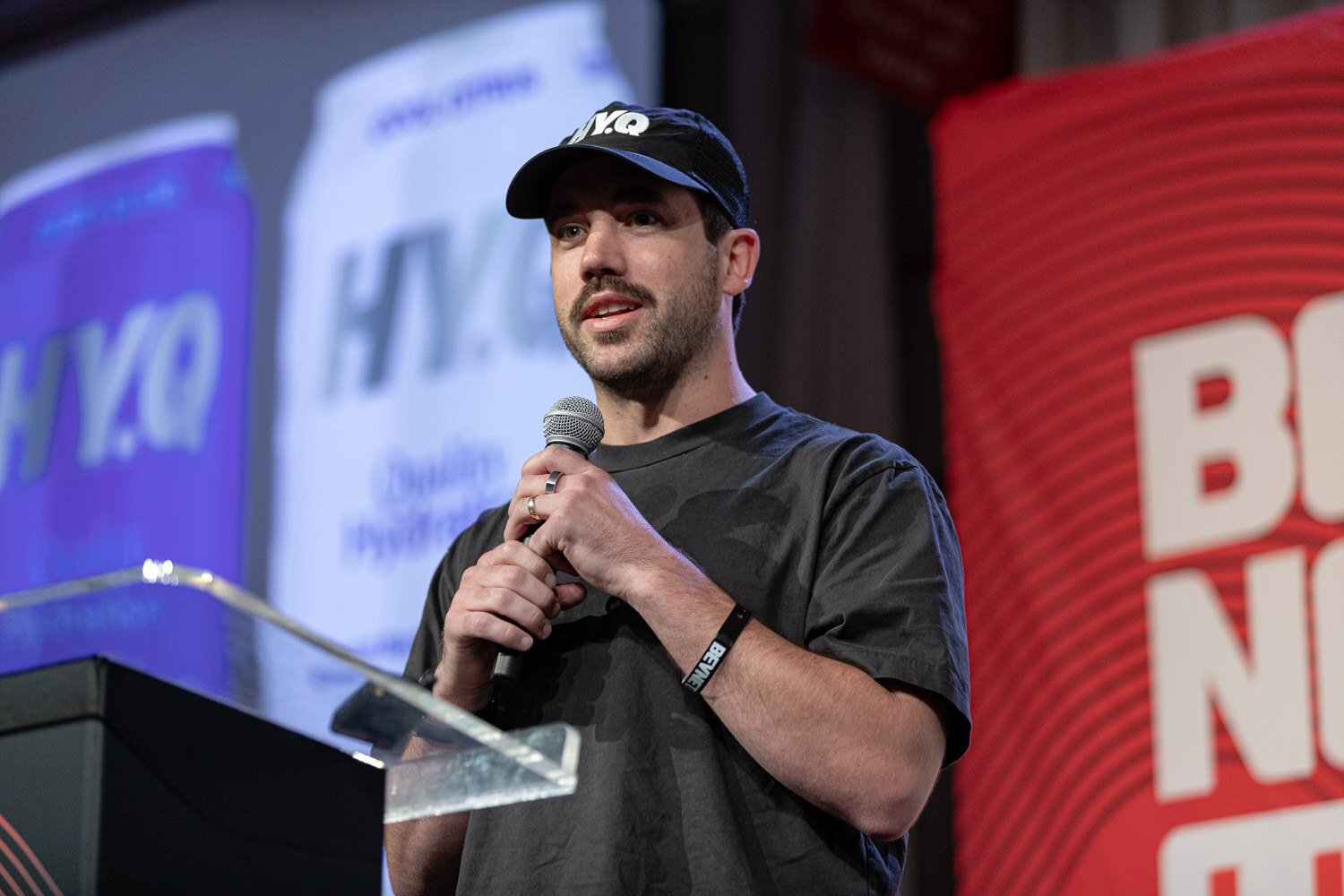 A man in a black HY.Q cap and gray t-shirt holds a microphone in front of a screen showing purple and white HY.Q cans.
