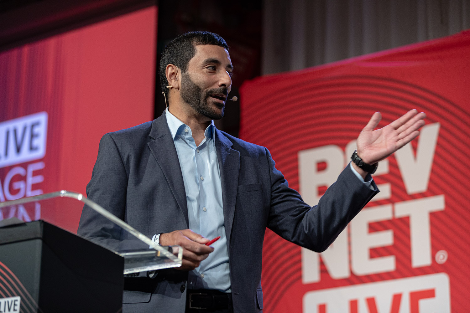 A man in a gray blazer holds a red marker and gestures beside a podium in front of a red BevNET banner.