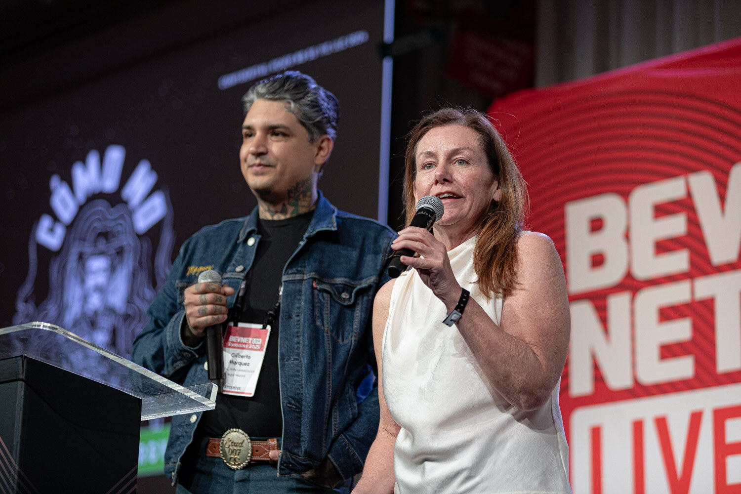 A man in a denim jacket and a woman in a white top each hold a microphone in front of a Cómo No slide.