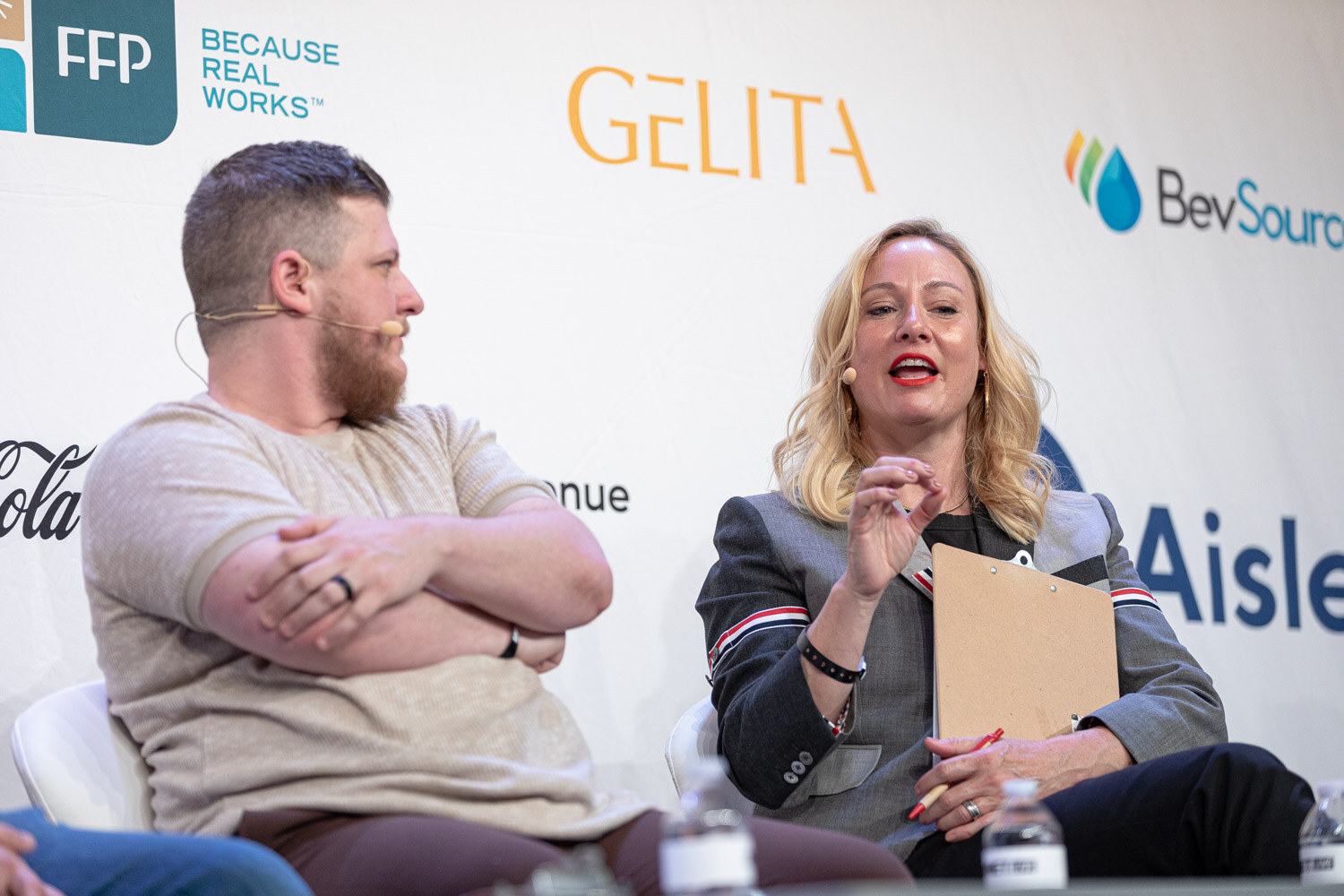 A bearded man in a beige t-shirt with crossed arms sits beside a woman with blonde hair and a clipboard.